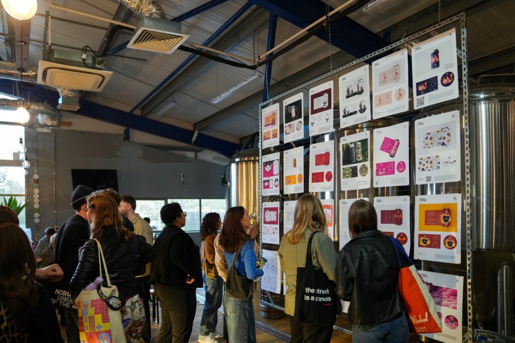 A group of people in a brightly lit room look at colorful prints and artwork displayed on a large wall grid during an exhibition or art event. Some carry tote bags and converse with each other.
