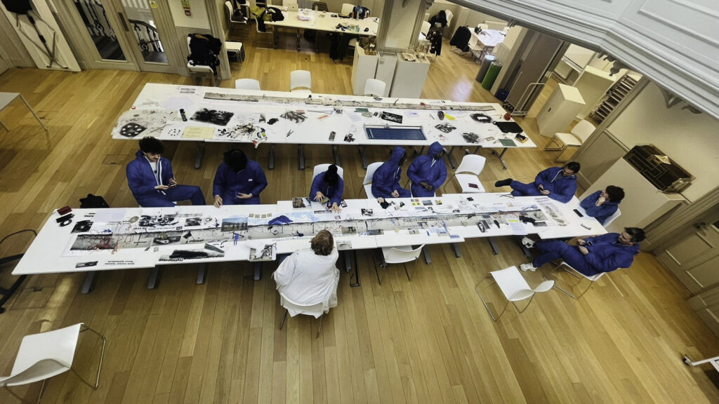 A group of people in blue coveralls sit around a long, white table covered with papers, tools, and art supplies in a spacious, well-lit room with wooden floors. One person in white sits at the head of the table.