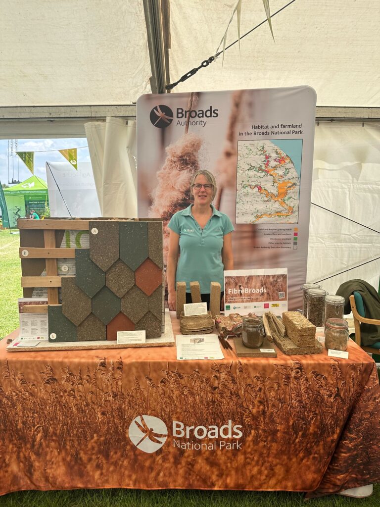 A woman stands at a Broads National Park display booth featuring paludiculture building materials, jars of samples, informational pamphlets, and a map poster inside a tent at an outdoor event.
