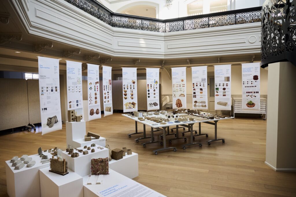 An exhibition room with wooden floors displays ceramic and architectural models on white pedestals and tables. Behind them, tall panels feature images and diagrams about paludiculture, with ornate balconies above and soft natural lighting.