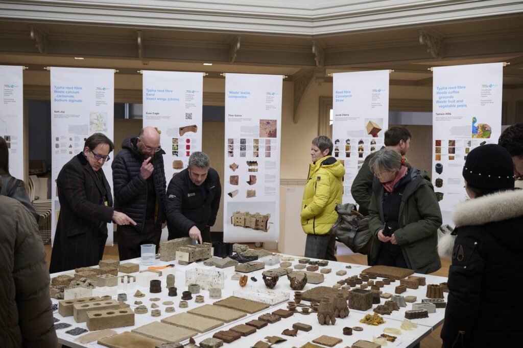 A group of people examines and discusses various brown building material samples displayed on tables in an exhibition space, with informational posters in the background.
