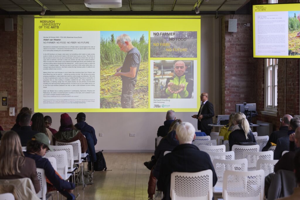 A speaker presents to an audience in a modern lecture room. A large screen displays slides about farming and paludiculture, including a man examining crops and the slogan: "No farmer, no food, no fibre, no future.