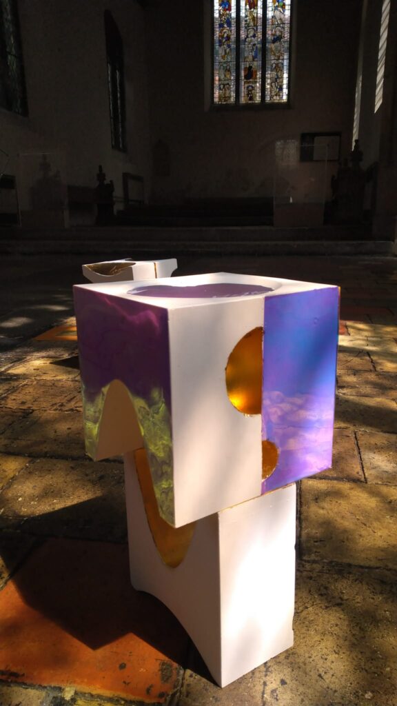A modern, geometric white sculpture with gold and purple accents stands on a sunlit stone floor inside a church, with stained glass windows and pews visible in the background.