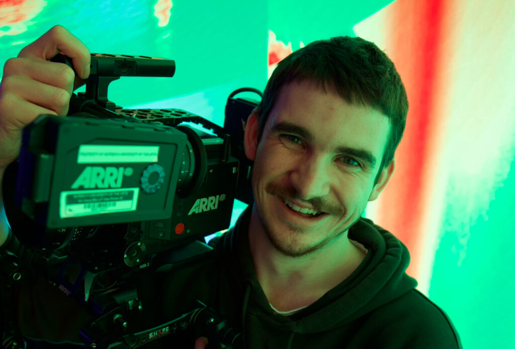 A smiling man holds a large ARRI video camera on his shoulder, standing in front of a brightly lit, colorful green and red background. He wears a dark hoodie and has a tattoo on his forearm.