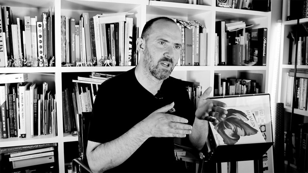 A man with a beard sits in front of a bookshelf filled with books, gesturing with his hands while speaking. A framed picture with an image of a face rests on the shelf beside him.
