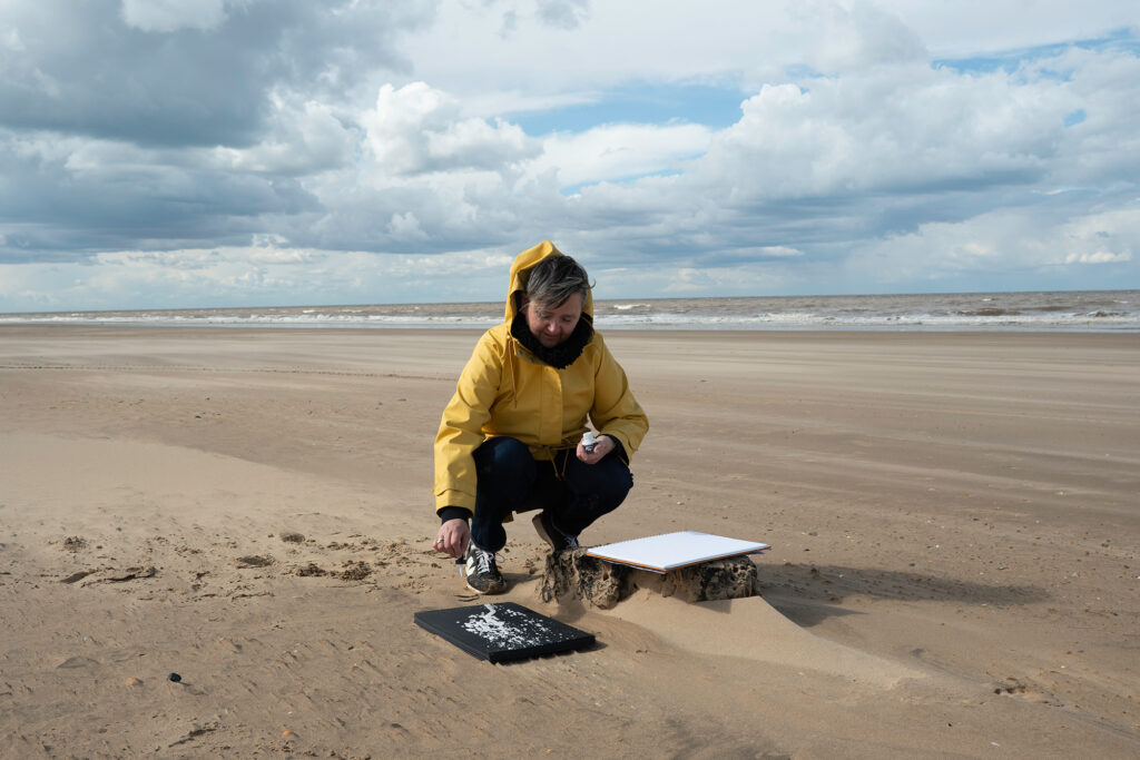 A person in a yellow jacket crouches on a sandy beach, arranging white shells or objects on a black board. A white board rests on a wooden block nearby, with the sea and cloudy sky in the background.