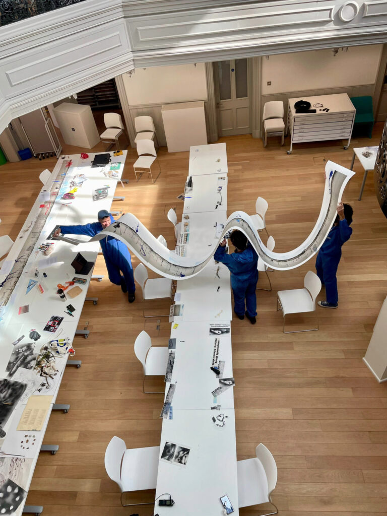 Two people in blue coveralls stand in a spacious, well-lit room, holding up a long, wavy sheet of paper that stretches across several white tables arranged end to end. Art supplies and papers are scattered on the tables.