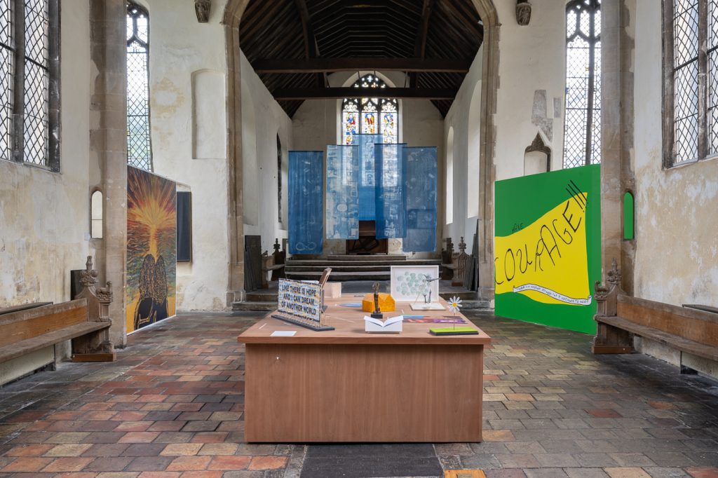 An art installation inside a church features colorful panels, including one with “COURAGE” in bold letters, drawings on a central table, and hanging blue fabric with stained glass windows in the background.Damian Griffiths, courtesy of Hospital Rooms