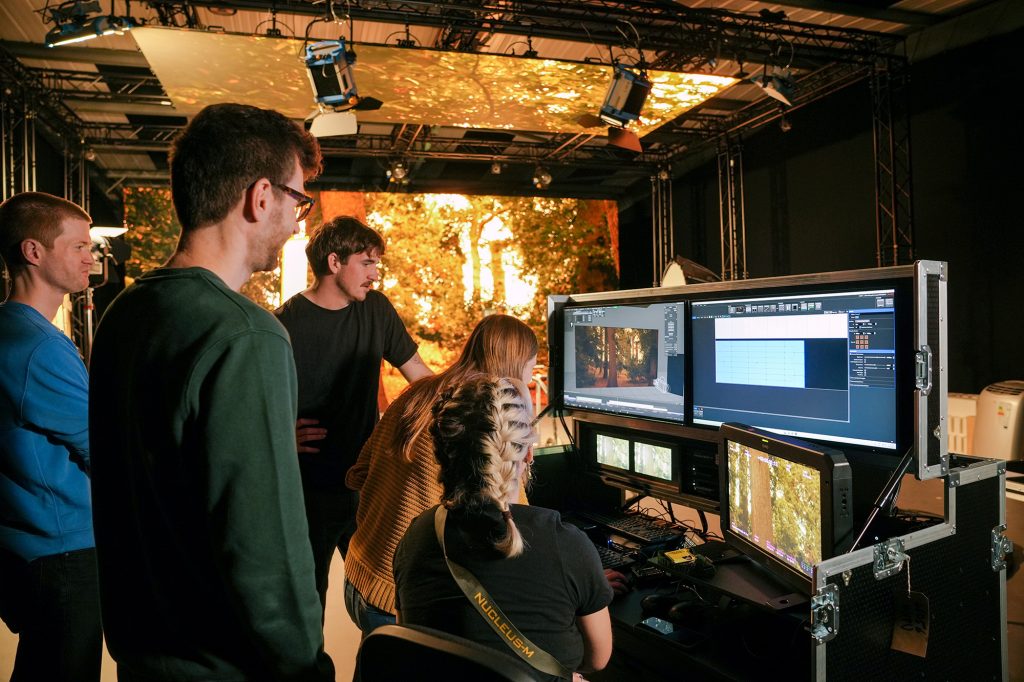 A group of people work together in a studio, gathered around computer monitors displaying digital graphics and a forest scene, with cables and equipment visible in the background.