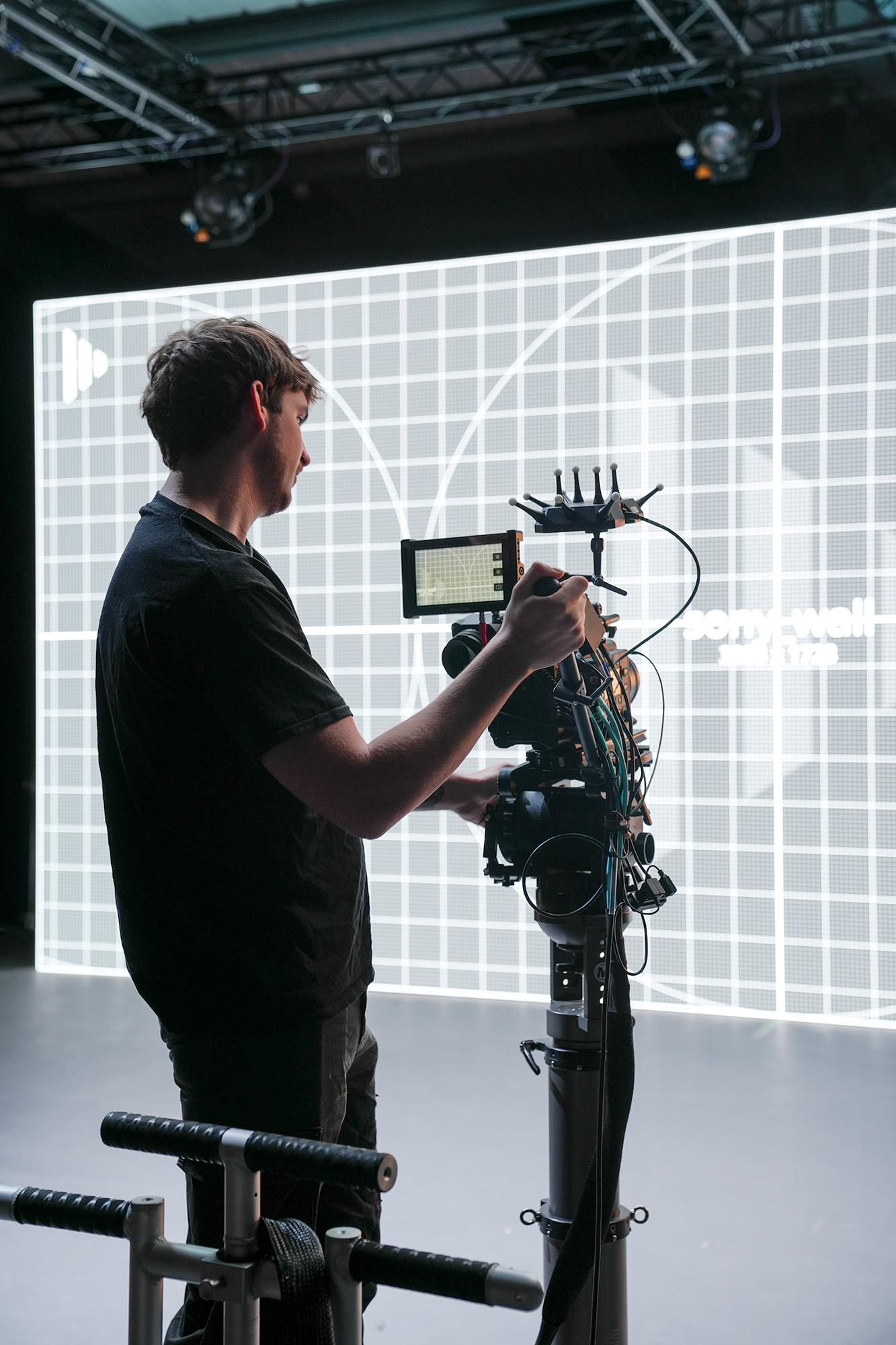 A person operates a professional video camera mounted on a rig in a studio, with a large grid-patterned screen and soft overhead lighting in the background.