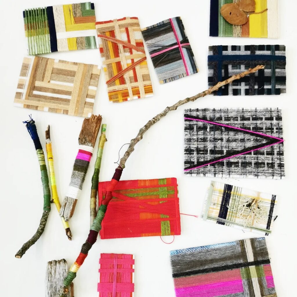 Various pieces of woven art, featuring colorful threads wrapped around cardboard and sticks, are arranged on a white surface. The designs include stripes, checks, and abstract patterns in vibrant colors.