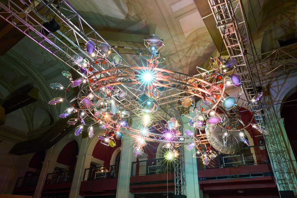 A large, circular, modern chandelier with colorful lights and reflective surfaces is suspended from a metal frame inside a grand hall with high ceilings and arched windows.