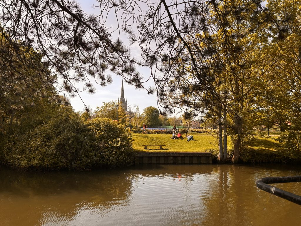 People relax on a grassy riverside under trees with autumn leaves, with Norwich Cathedral visible in the background across the water on a sunny day.
