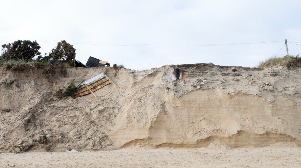 Eroded sandy cliff with debris from a collapsed structure, including wood and metal, hanging over the edge; some vegetation and a utility pole are visible at the top against a cloudy sky.
