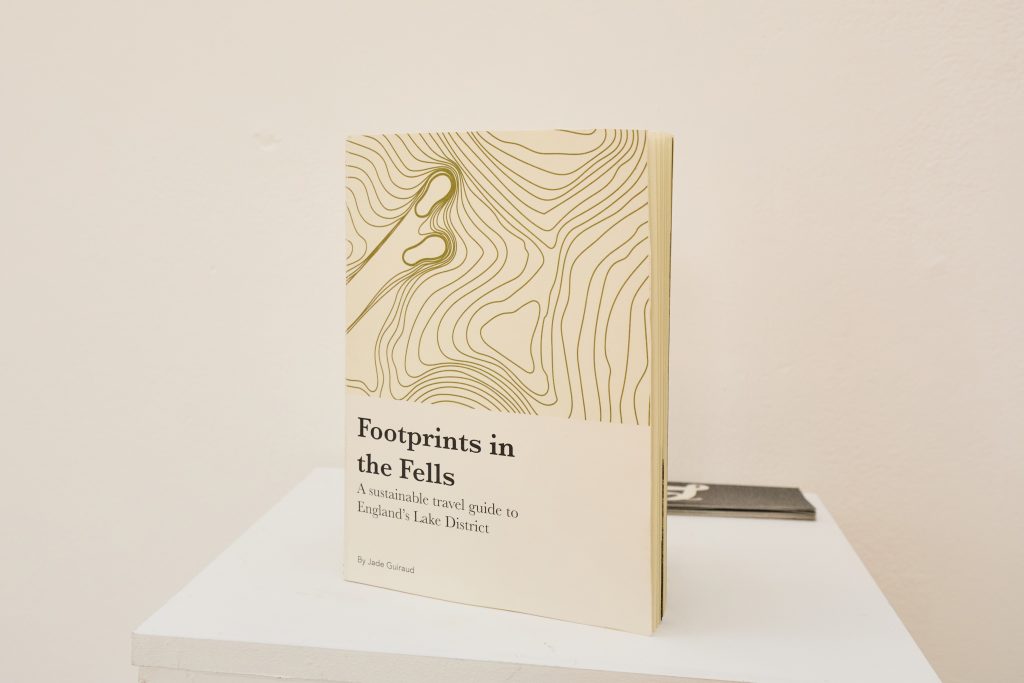 A book titled "Footprints in the Fells: A sustainable travel guide to England's Lake District" with a tan cover featuring topographic lines and a footprint, standing upright on a white plinth.