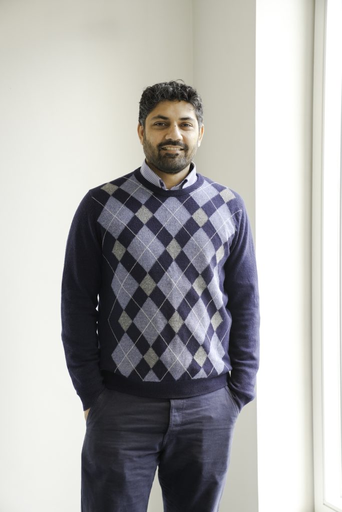 A man with dark curly hair and a beard stands by a bright window, wearing a blue argyle sweater over a collared shirt and dark trousers. He is smiling and has his hands in his pockets.