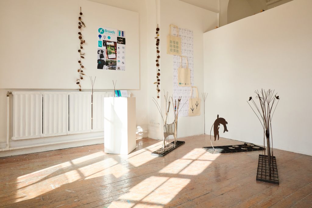 A bright art gallery space with wooden floors, featuring metal animal sculptures, wall hangings and posters, with sunlight streaming through large windows onto the floor.
