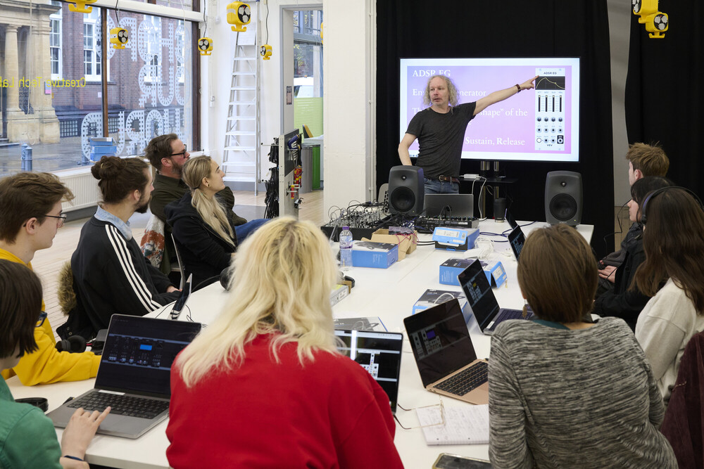 A group of people with laptops sit around a table in a bright classroom, listening to a presenter who is pointing at a projected screen with audio editing software displayed.