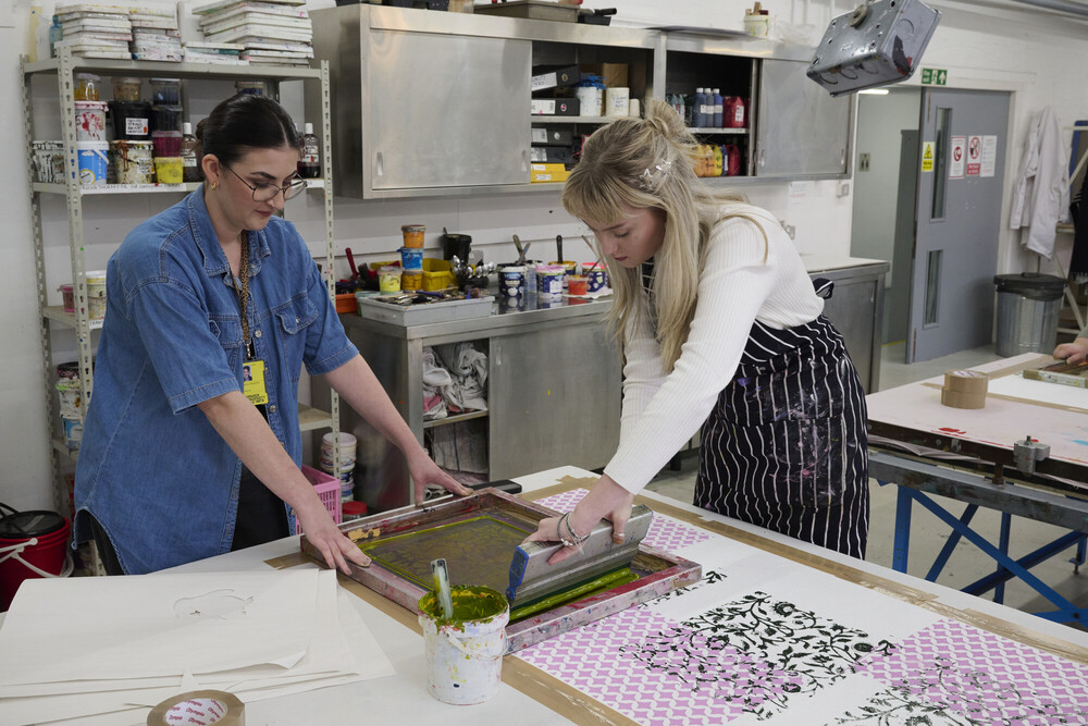 Two women work together on a screen printing project in an art studio. One holds the frame steady while the other pulls ink across the screen. Art supplies and colorful prints are visible on the table and shelves.