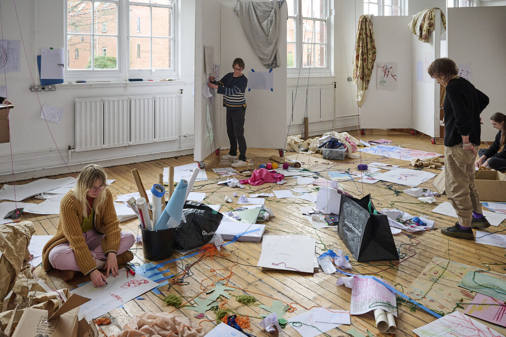 Four people work on art projects in a bright, messy studio scattered with papers, drawings, and art supplies. Large windows let in natural light, and artworks hang on the walls and floor.