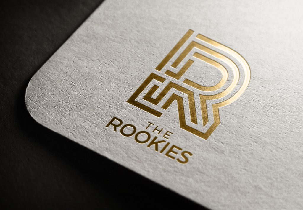 A close-up of a textured card featuring a gold, geometric "R" logo above the words "THE ROOKIES" in elegant, capital letters. The design and text are embossed and metallic.