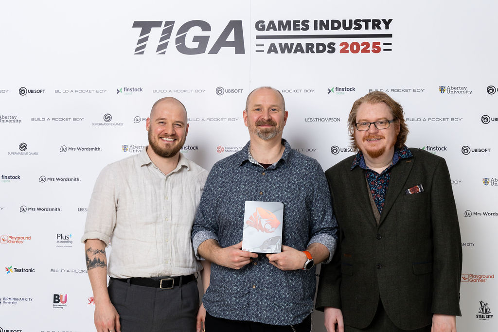 Three people are standing in front of a white wall with 'TIGA' written in bold print behind them. The person in the middle, who wears a blue shirt, holds a trophy.