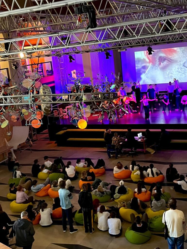 A group of people sits on colorful bean bags watching a stage performance with lights and a large projection screen, while some participants work with equipment and instruments onstage.