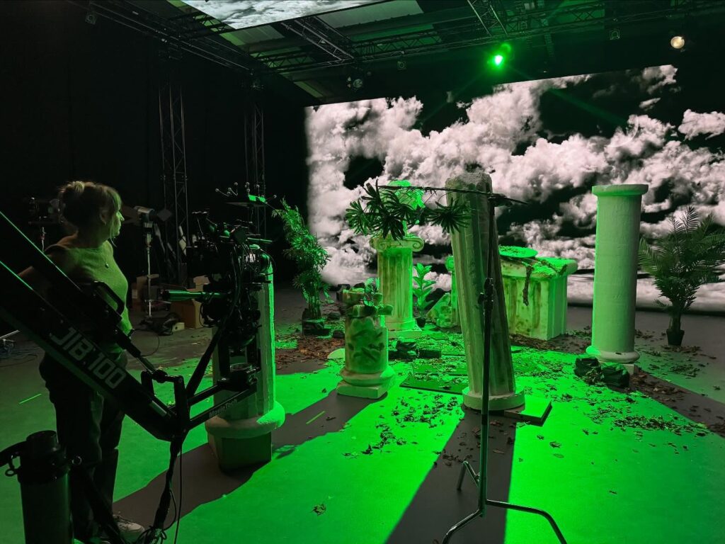 A film set with white ancient Greek-style columns, plants, and scattered leaves, lit by green light. A large screen displays cloudy skies, and a camera operator stands by a camera rig on the left.