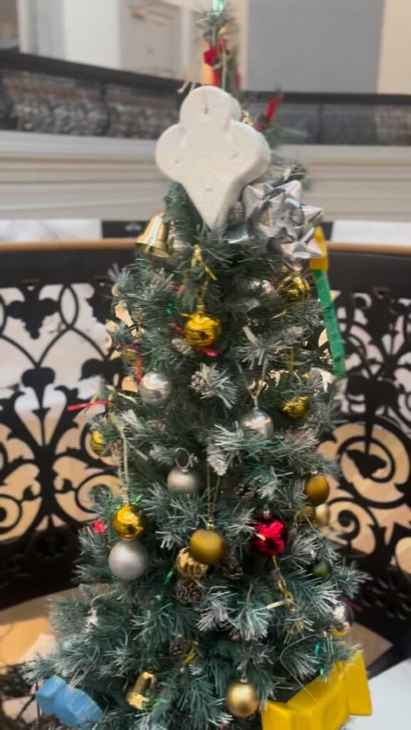 A small decorated Christmas tree with various ornaments, including gold and silver balls, a metallic bow, and a white star-shaped topper, with a few wrapped presents at the base. The tree is set against an ornate black railing.