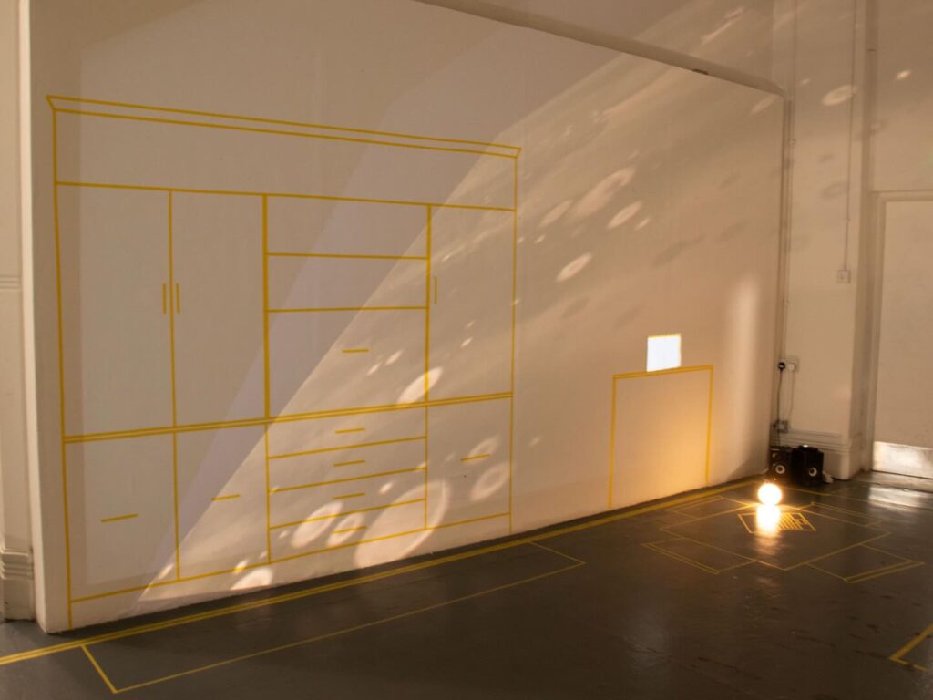 A room with a wall decorated with yellow tape outlines resembling cabinets and drawers. Sunlight reflects circular patterns on the wall and floor. A small lit bulb sits on the floor near a cut-out window.