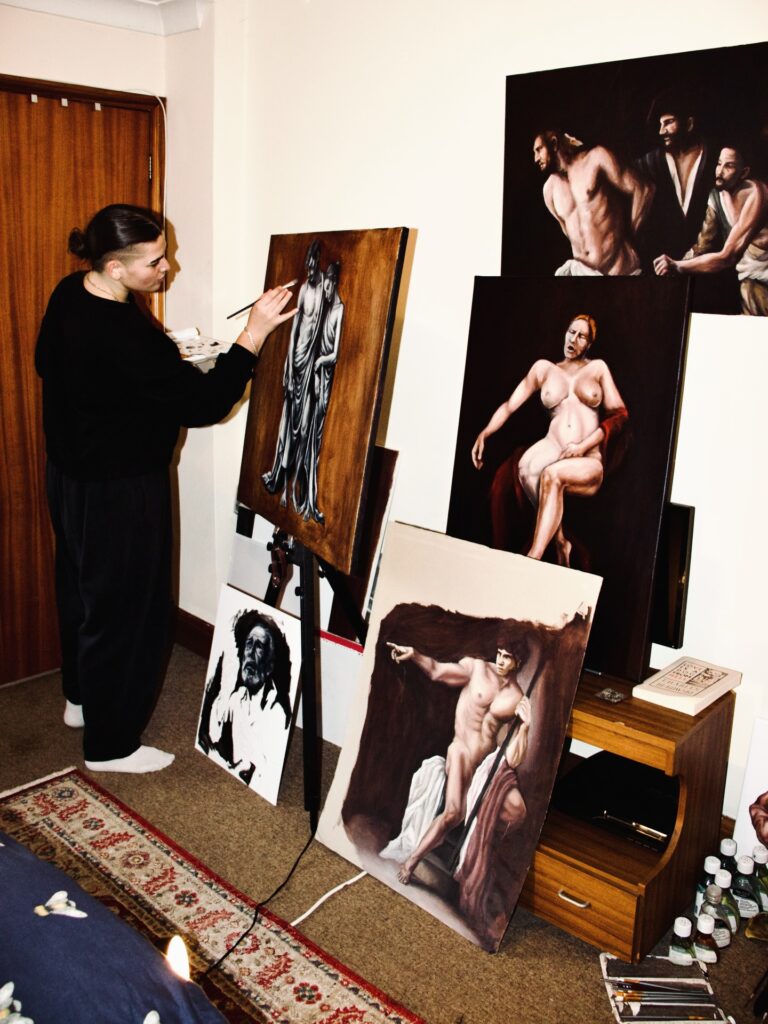A person paints a classical-style portrait on an easel in a room filled with several other realistic figure paintings leaning against the wall and furniture. The space has warm lighting and a cozy, creative atmosphere.