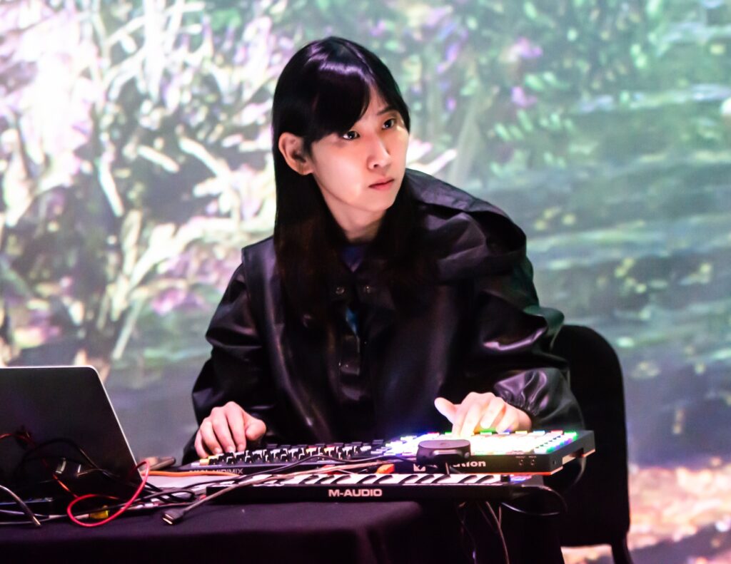 A person with long dark hair operates electronic music equipment and a laptop, sitting at a table in front of a colorful, out-of-focus background that resembles a nature scene.