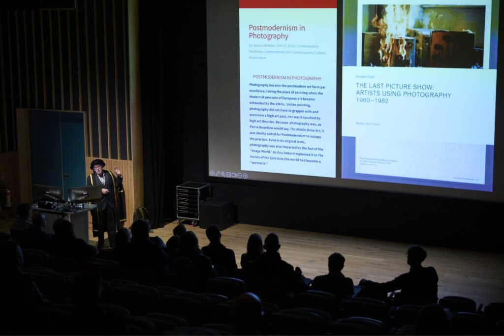 A person in dark clothing presents a lecture on postmodernism in photography to an audience in a dimly lit theater, with slides projected on a large screen at the front of the room.