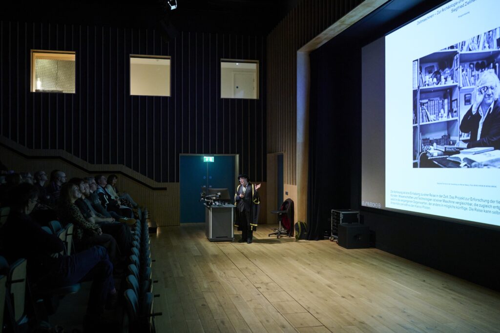 A person gives a presentation in a dark auditorium, standing at a podium in front of an audience. A large projected slide displays a black and white photo and text on the screen beside them.