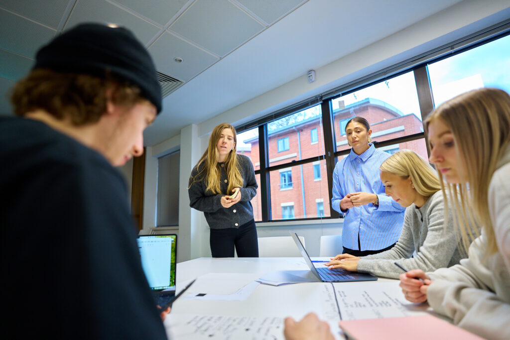 Three students sit at a white table with laptops and notes in a bright room. Behind them, two lecturers stand in front of large windows, showing a red brick building outside.