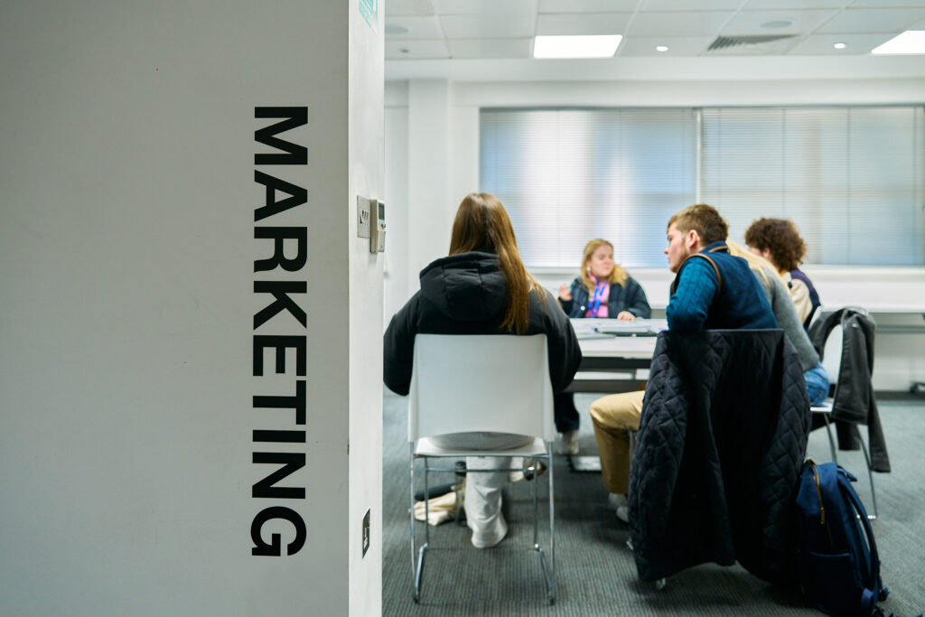 Four people sit around a table in a modern university room. The word "MARKETING" is prominently displayed on a wall in the foreground.