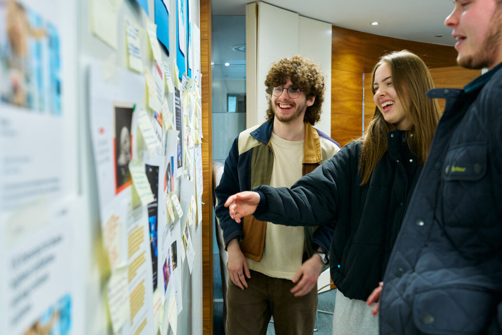 Three students stand together, smiling and laughing as they look at and point to notes and posters pinned on a bulletin board in an indoor, modern setting.