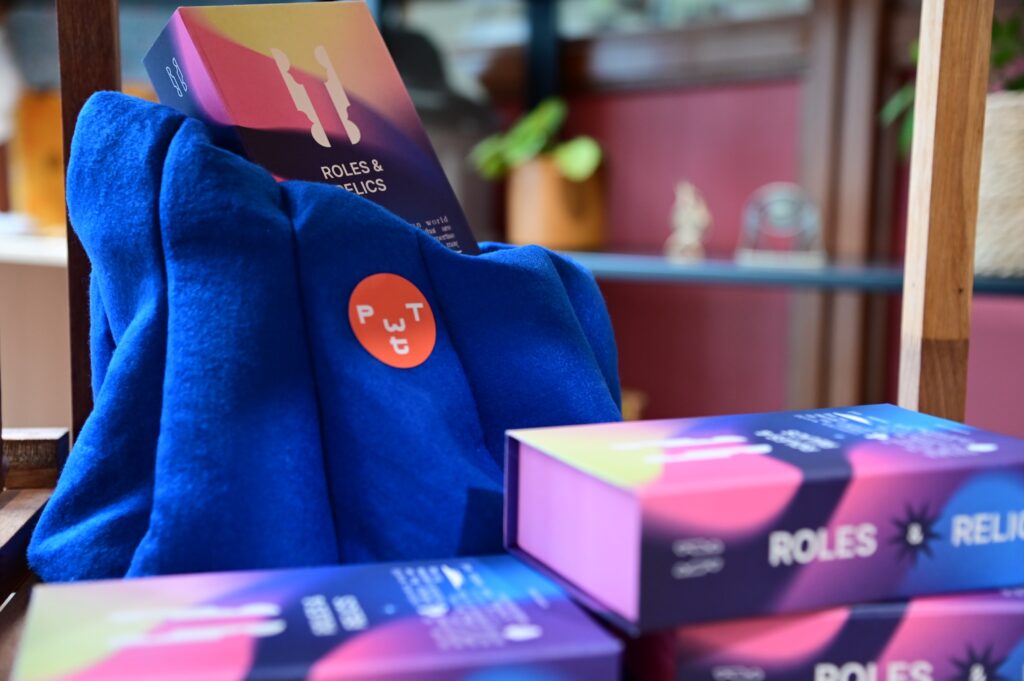 A stack of colorful game boxes labeled "Roles & Relics" sits on a chair draped with a bright blue fabric and a round orange badge with the letters "PWT." Blurred indoor plants and shelves are in the background.