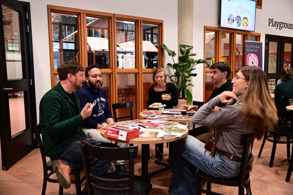 Five people sit around a circular table in a modern indoor space, talking and playing a board game. Magazines and game pieces are spread out on the table. There are large windows and potted plants in the background.