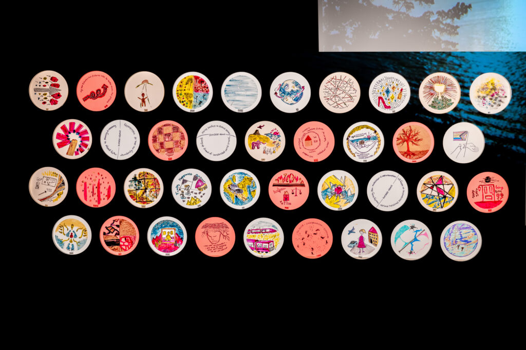 A grid of 36 round coasters, each featuring a unique, colorful drawing or design, is displayed on a black background. A blue-toned photo with leaves appears in the top right corner.