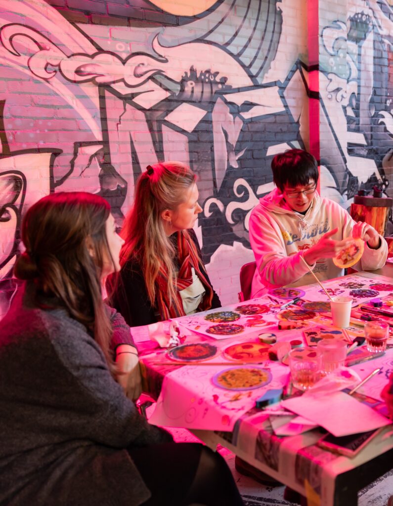 Three people sit around a table covered with art supplies and colorful materials, talking and working on crafts together. A wall behind them is decorated with graffiti-style art. Warm lighting fills the scene.