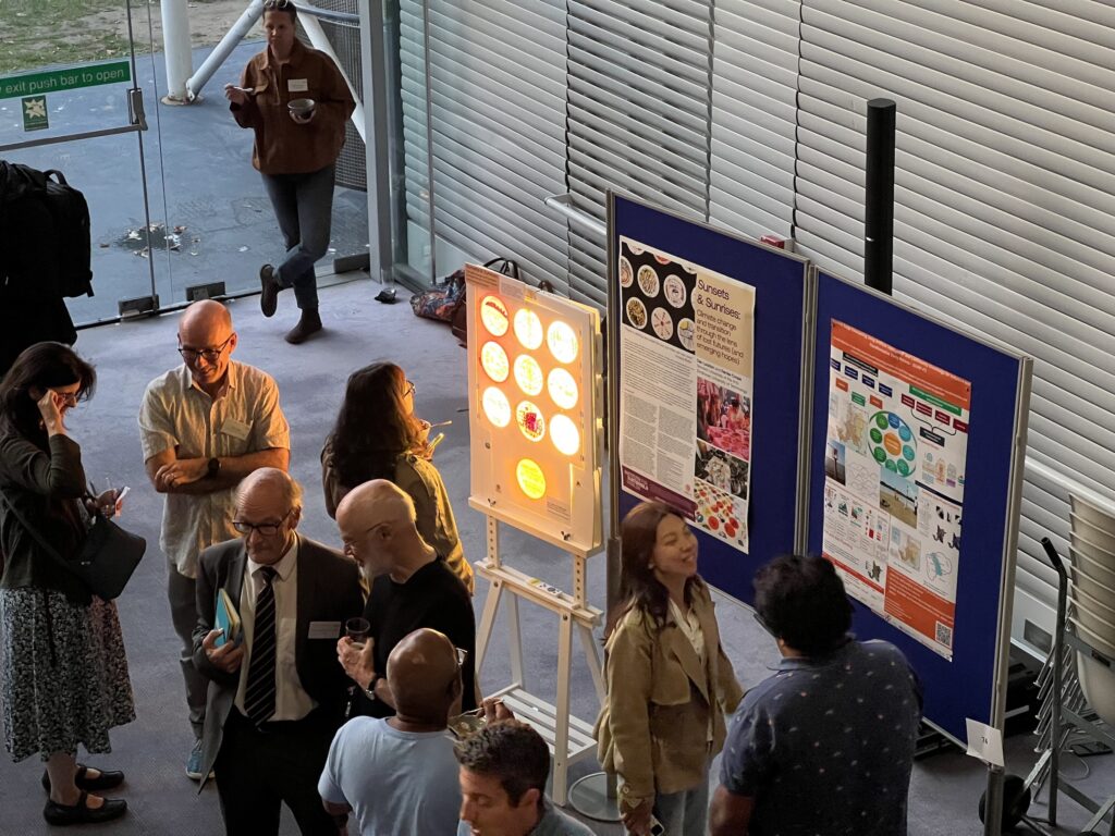 Overhead view of people conversing at an indoor event near posters and a brightly lit display board, with windows and an exit door visible in the background.