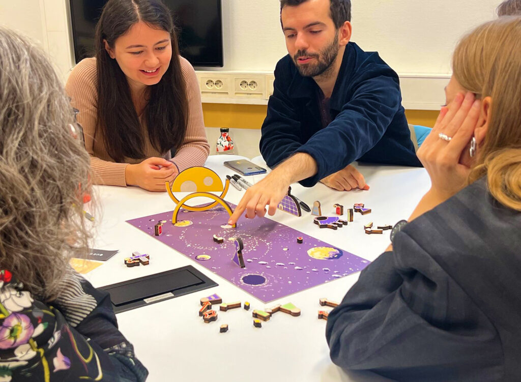 Four people sit around a table, engaged with a colorful board game featuring small pieces and cards. One man points to the board while others watch and discuss, creating an atmosphere of focus and collaboration.