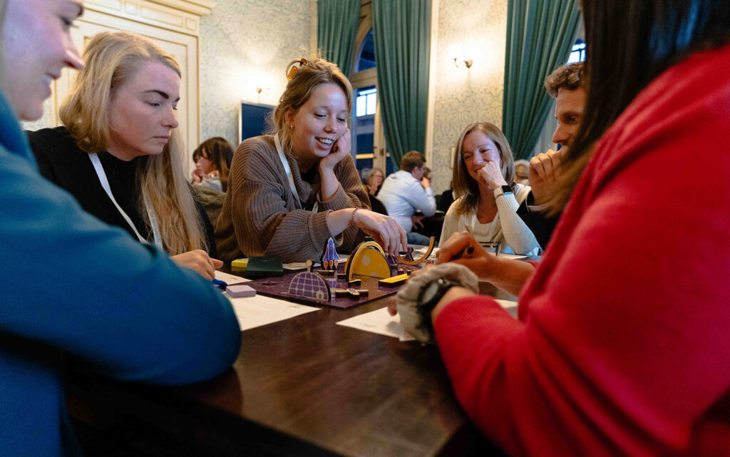 A group of people sit around a table, smiling and talking while playing a board game together in a warmly lit room with elegant décor and large curtains.