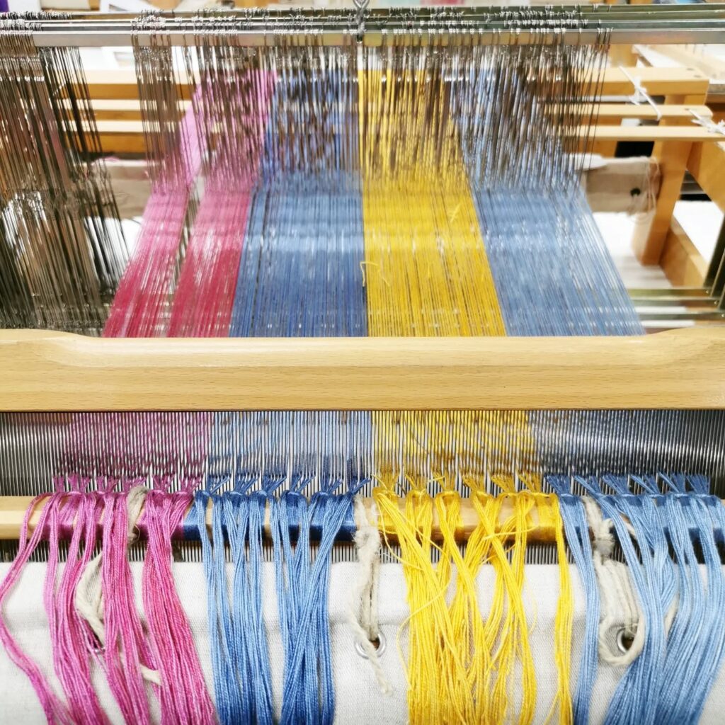 Close-up of a weaving loom with colorful threads in pink, yellow, and blue, arranged neatly and stretched through the loom’s heddles and reed, ready for weaving.