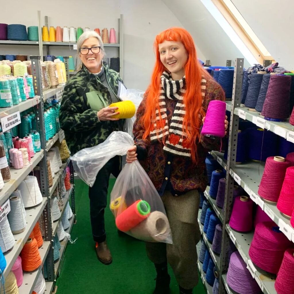 Two people stand among shelves filled with colorful spools of yarn. One holds a yellow spool, and the other carries a bag filled with yarn. Both are smiling. The setting appears to be a yarn or craft supply store.