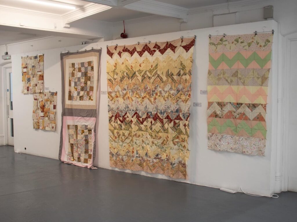 Four colorful patchwork quilts with various geometric patterns hang on a white wall in an art gallery with a gray floor and white ceiling. Each quilt features different shapes and pastel shades.