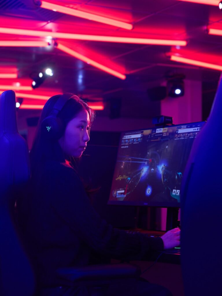A woman wearing headphones intently plays a video game on a computer in a neon-lit gaming room with pink and purple lights overhead.