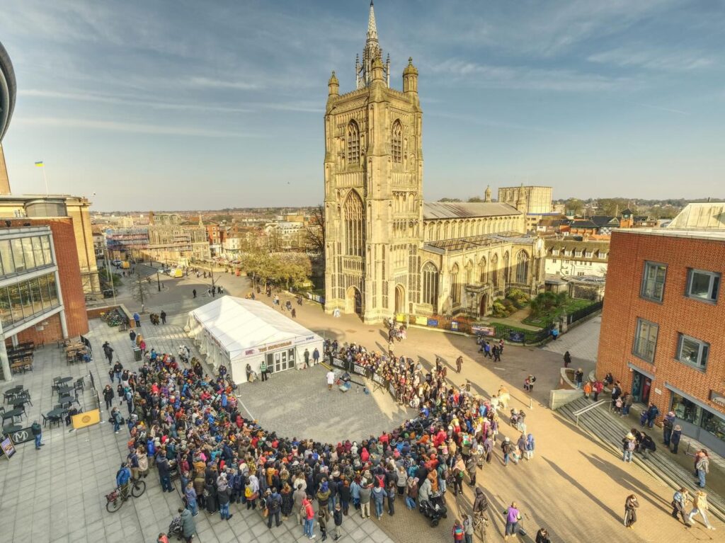 A large crowd gathers in a city square outside a historic stone church with a tall tower, surrounded by modern and traditional buildings under a clear sky.