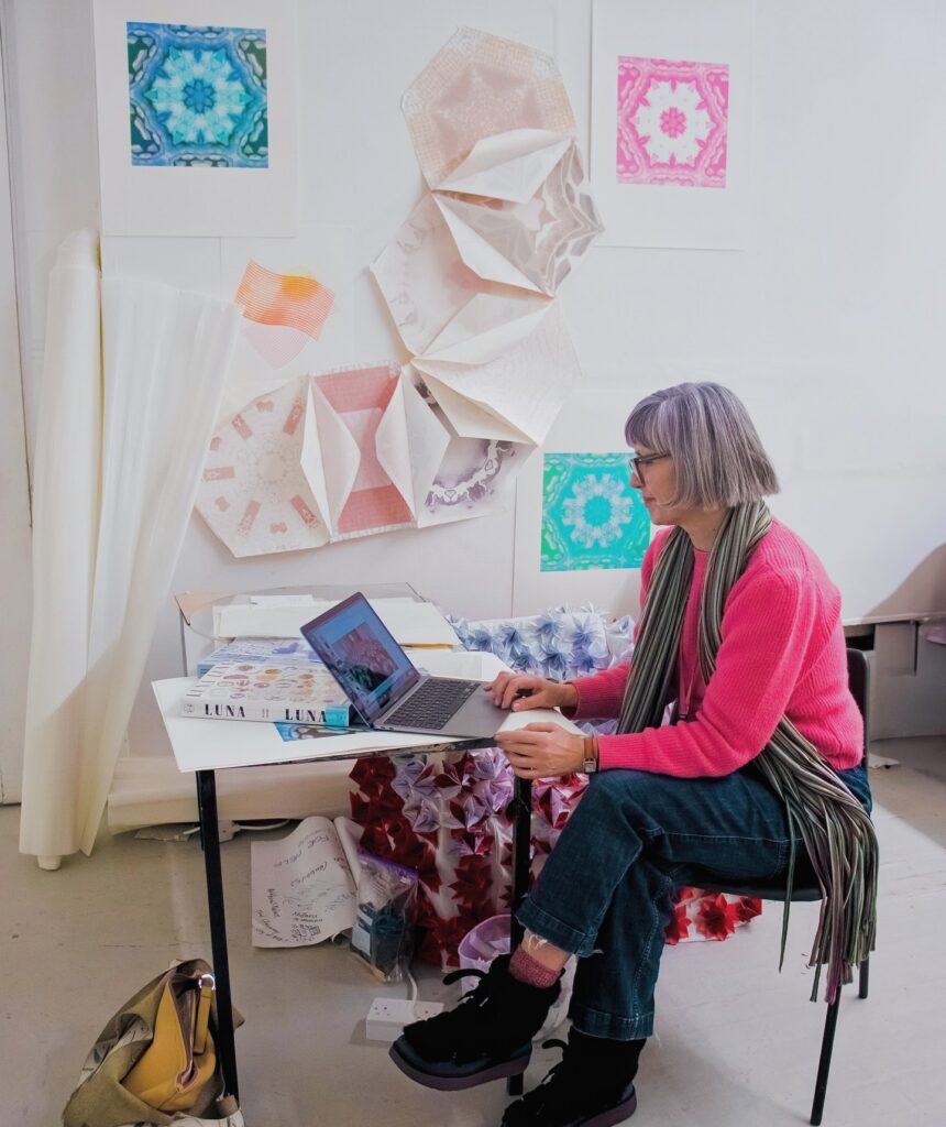 A woman with gray hair and glasses, wearing a pink sweater and scarf, sits at a desk using a laptop. Colorful geometric art and fabric samples are displayed on the white wall behind her.